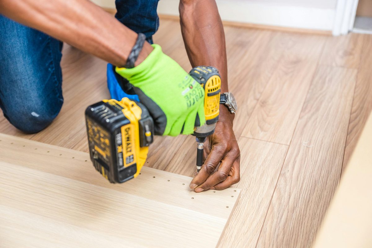 Hands in a lime-green work glove using a cordless drill to assemble wooden furniture — illustrative of Jax Ops hands-on craftsmanship